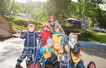 Fem barn på trehjulssykler en solrik sommerdag med grønt gress og høye trær i Fjellvegen barnehage