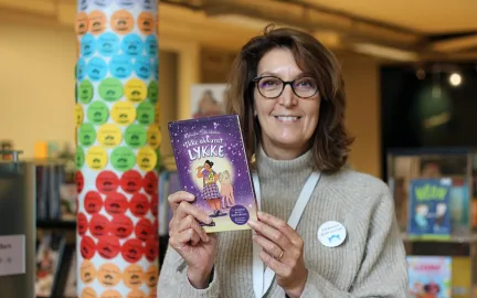 Lesetips-sommerles En bibliotekar smiler og holder opp boka "Ikke akkurat lykke"