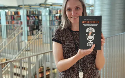 Boktips-Røverhistorier Dame med skulderlangt hår og mønstrete bluse holder boka Røverhistorier og smiler mot kamera. Foto.