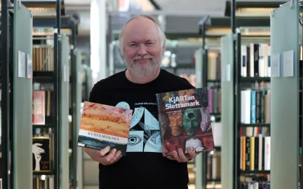 Øystein 150-årsutstilling Mann i svart skjorte med lyseblått trykk holder opp to bøker og smiler mot kamera. Foto.