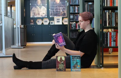 Regine_dødsforelska Kvinne sitter på gulvet og leser ei bok med ryggen lent mot ei bokhylle. Hun er kledd i svart og har lange, svarte støvletter på seg. Foran henne står to bøker med fronten mot kamera. Foto.