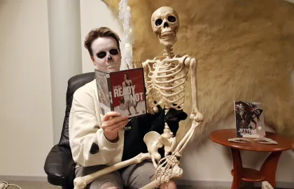 Kvinne med Halloweensminke i ansiktet sitter med et leke-skjelett p&aring; fanget. Hun holder opp et filmdeksel. Foto