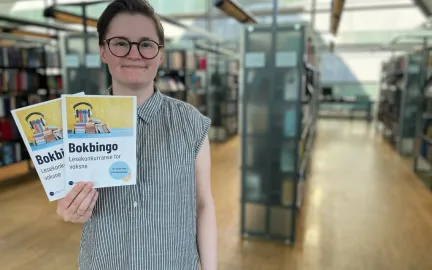 Lesetips til bokbingo Kvinne med kort brunt hår, briller og grå-og-hvit-stripete skjorte holder to ark og smiler mot kamera. I bakgrunnen er vinduer og bokhyller. Foto.