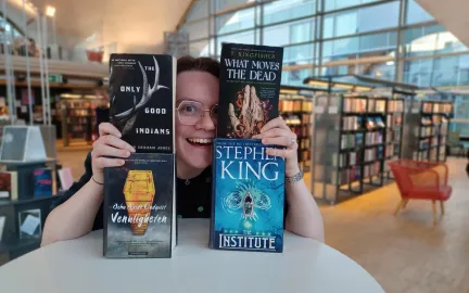 Oktober-lesetips 2024 Kvinne holder opp fire bøker og smiler bredd. Ansiktet hennes stikker frem mellom bøkene. I bakgrunnen en stor glassvegg og bokreoler. Foto.